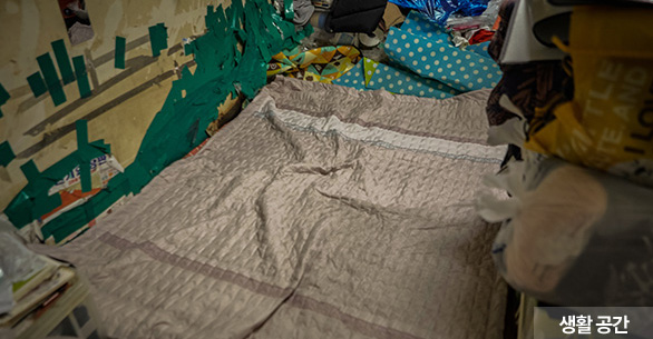 생활 공간, 방의 3분의 1을 제외한 곳에는 물건들이 쌓여 있고 우진이와 엄마가 잠을 자고, 먹고 모든 생활을 하는 공간에는 1인용 이불이 펴져있으며 벽에는 초록색 청테이프가 덕지덕지 붙여져 있는 모습
